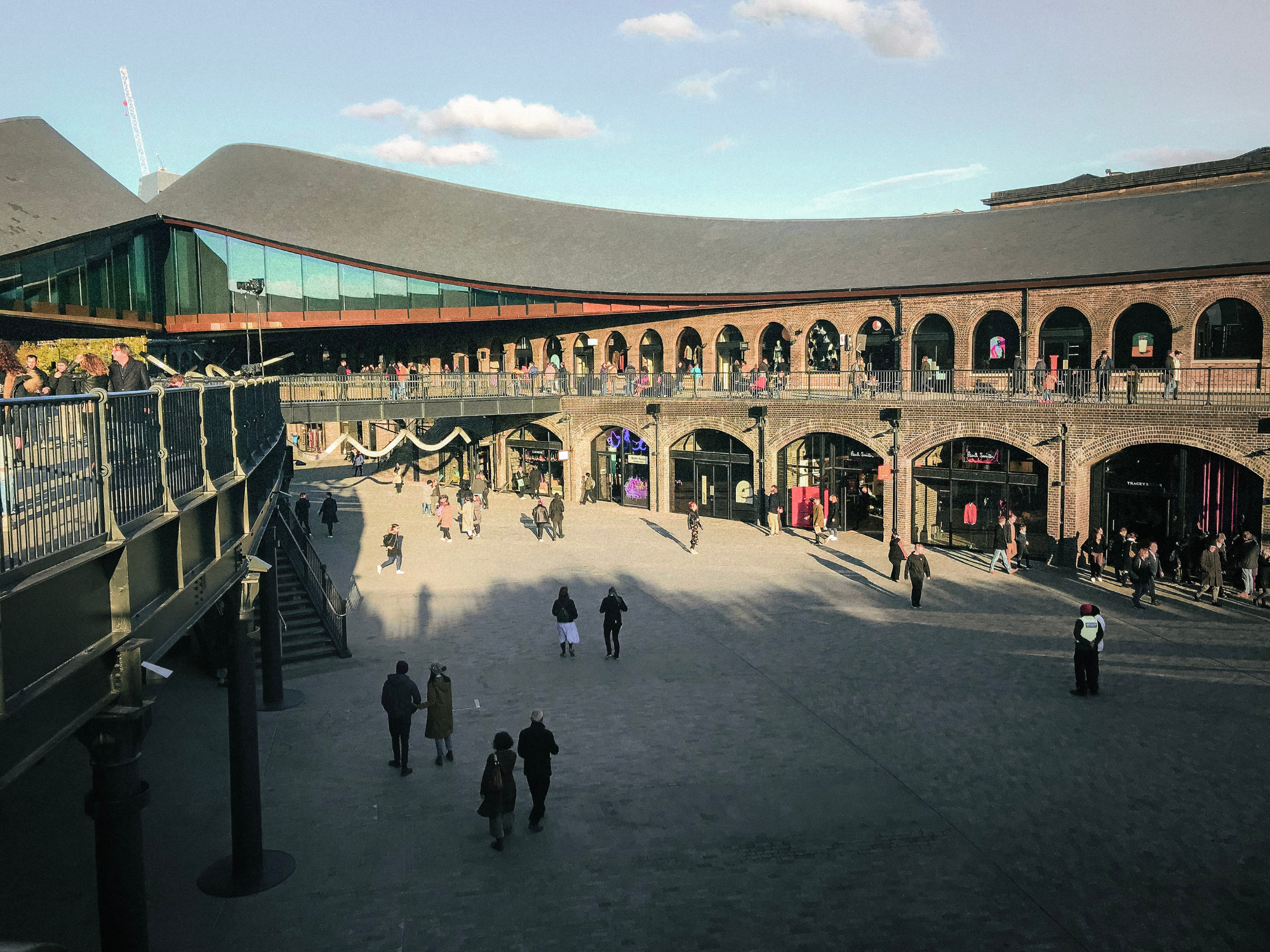Crowd of people walking in a brick courtyard with arched windows and a modern roof in London.