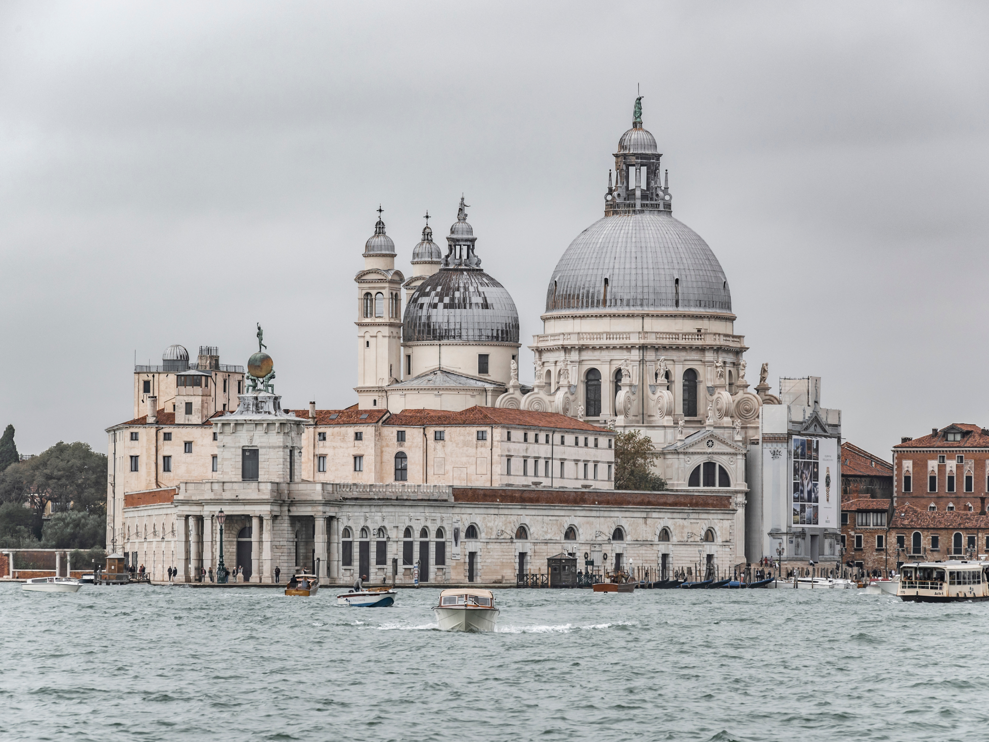 Historic buildings along the waterfront in Venice, featuring domes and ornate architecture under a cloudy sky.