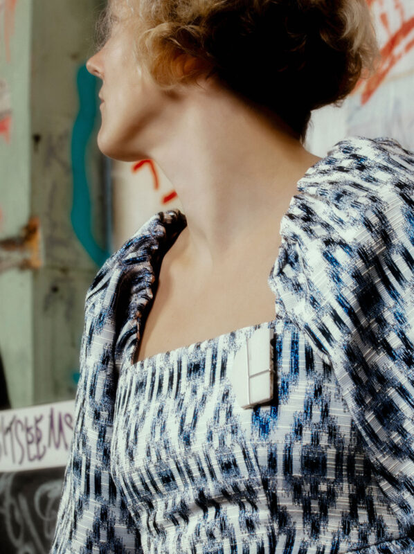 A person wearing a patterned dress with a brooch attached, posed against a graffiti-covered wall.
