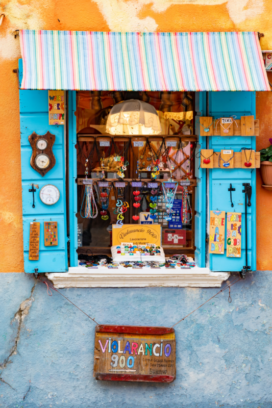 Colorful shop window in Venice displaying jewelry and souvenirs, with a striped awning and an orange wall backdrop.