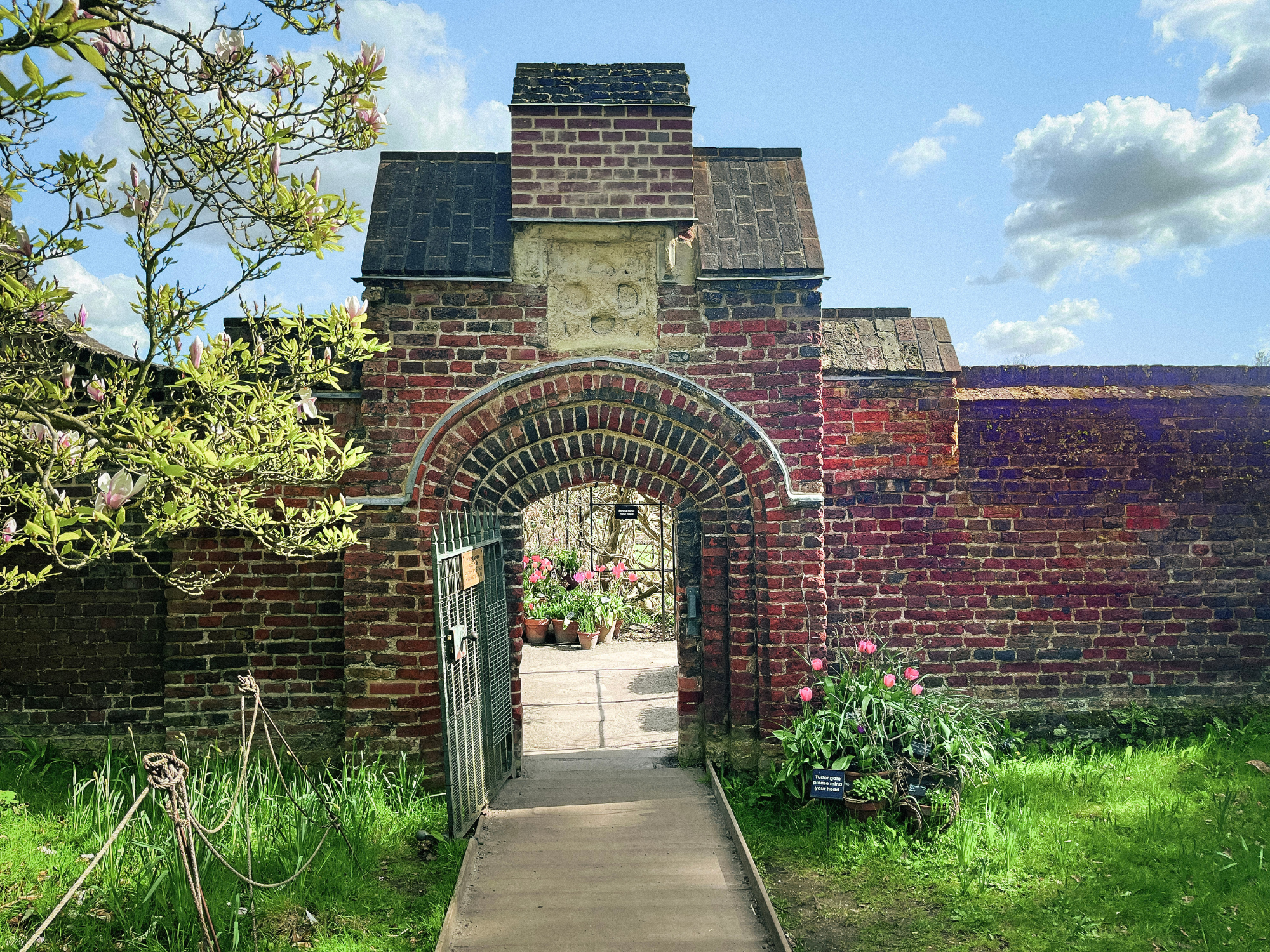 Brick archway leading to a garden, surrounded by greenery and flowers, under a blue sky with clouds.