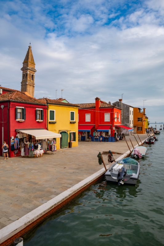 Colorful buildings line a canal in Burano, Venice, with boats docked and a bell tower in the background.