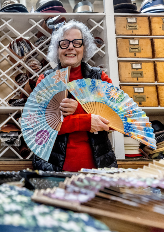 Woman with curly gray hair and glasses holds two colorful fans in a shop filled with hats and crafting materials.