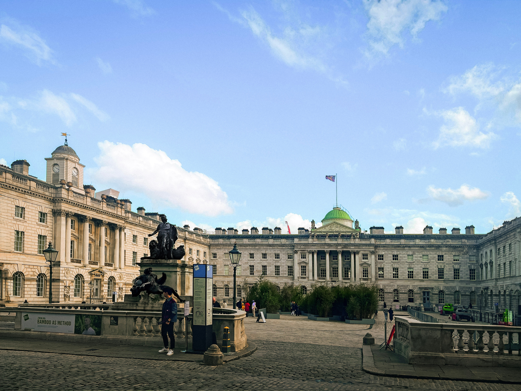 Somerset House courtyard in London, featuring a statue, green dome, and people enjoying the space under a blue sky.