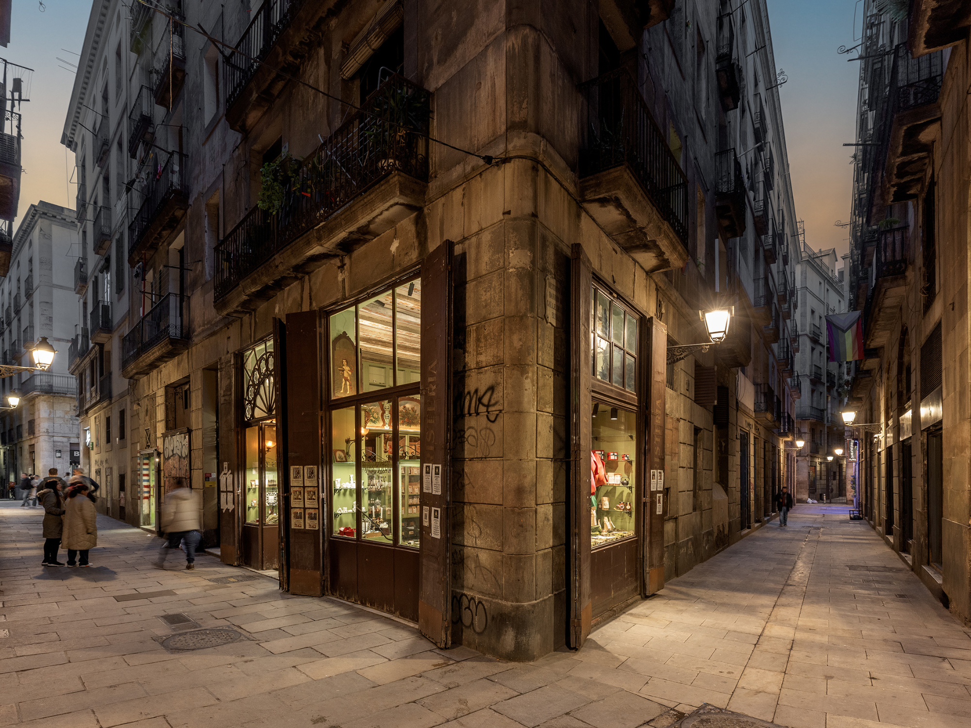 Narrow Barcelona street corner with shops, warm lighting, and people walking in the evening.