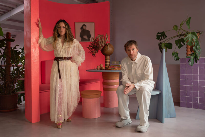 A woman in a sheer white dress and a man in a white outfit pose in a colorful interior with plants and modern furniture.