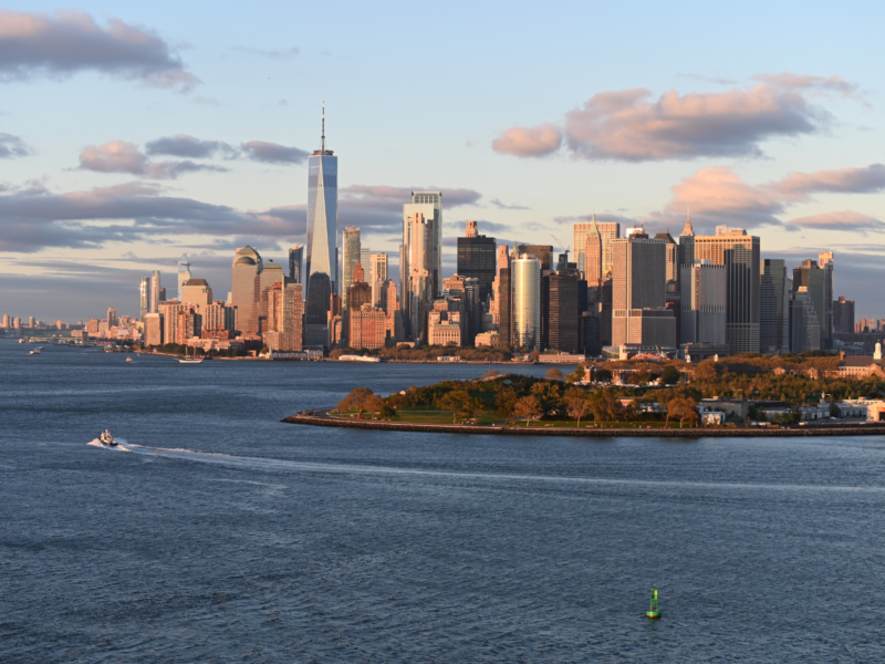 New York City skyline at sunset, featuring skyscrapers and a boat on the water, with a green buoy in the foreground.