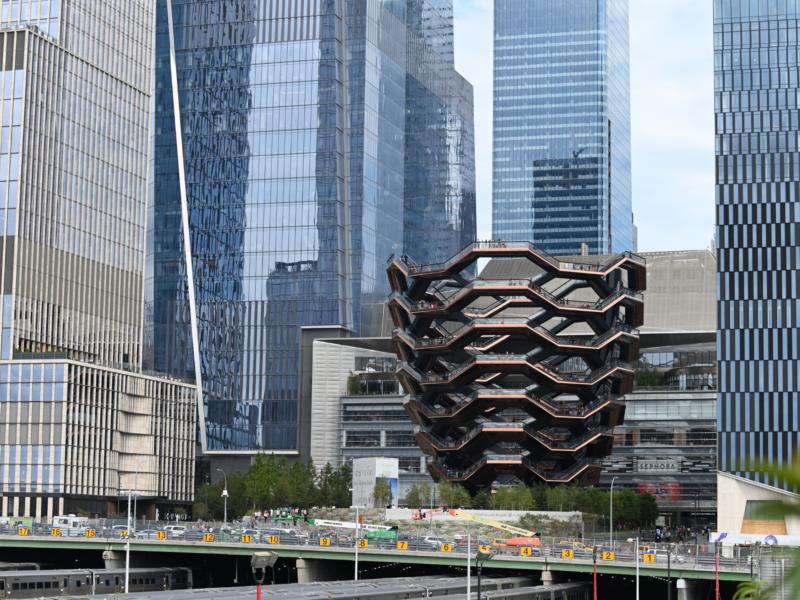 Vessel structure in Hudson Yards, surrounded by modern skyscrapers and greenery, with a busy street below.