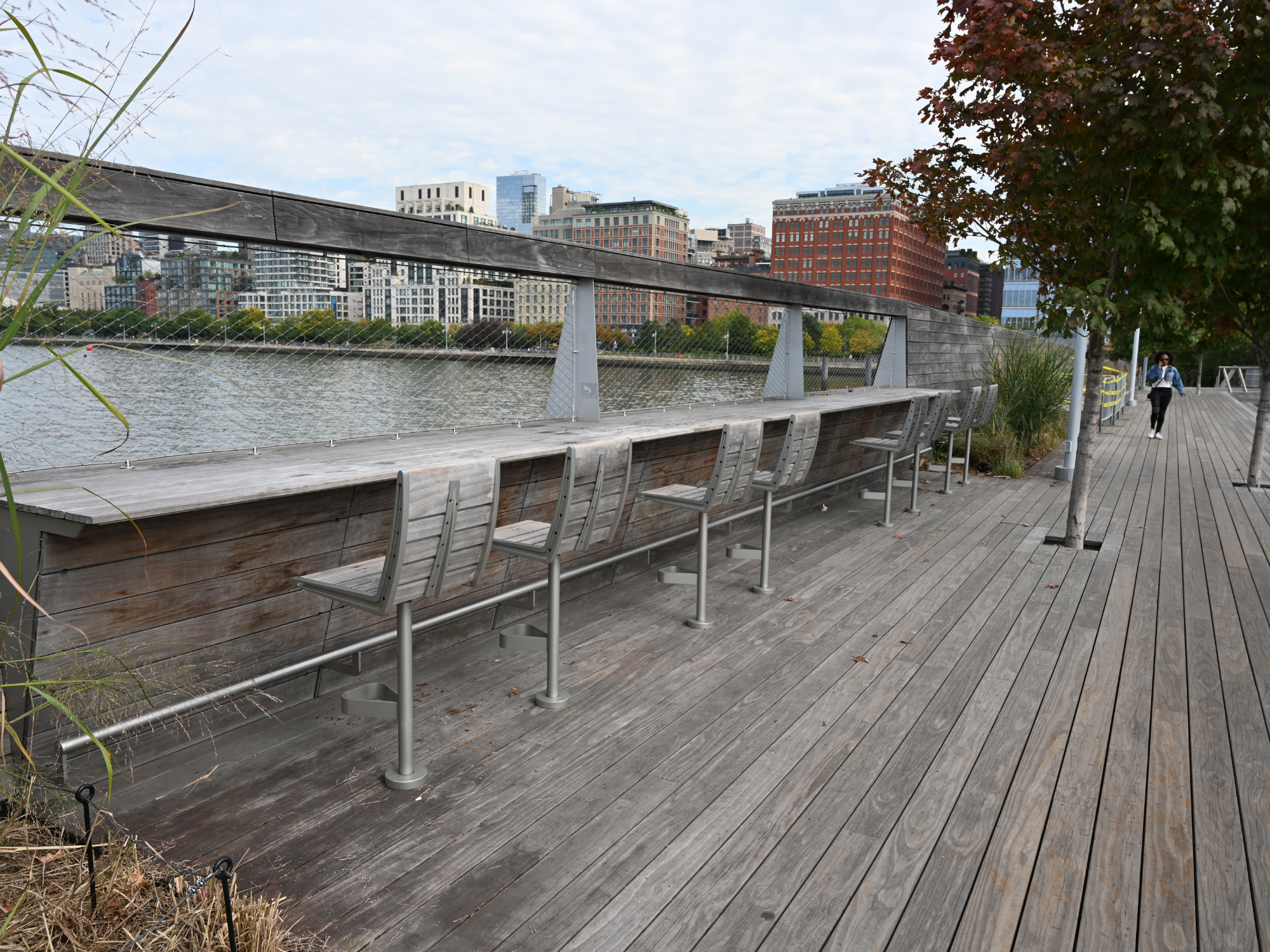 Wooden waterfront seating area with metal chairs, overlooking a river and city skyline under a cloudy sky.