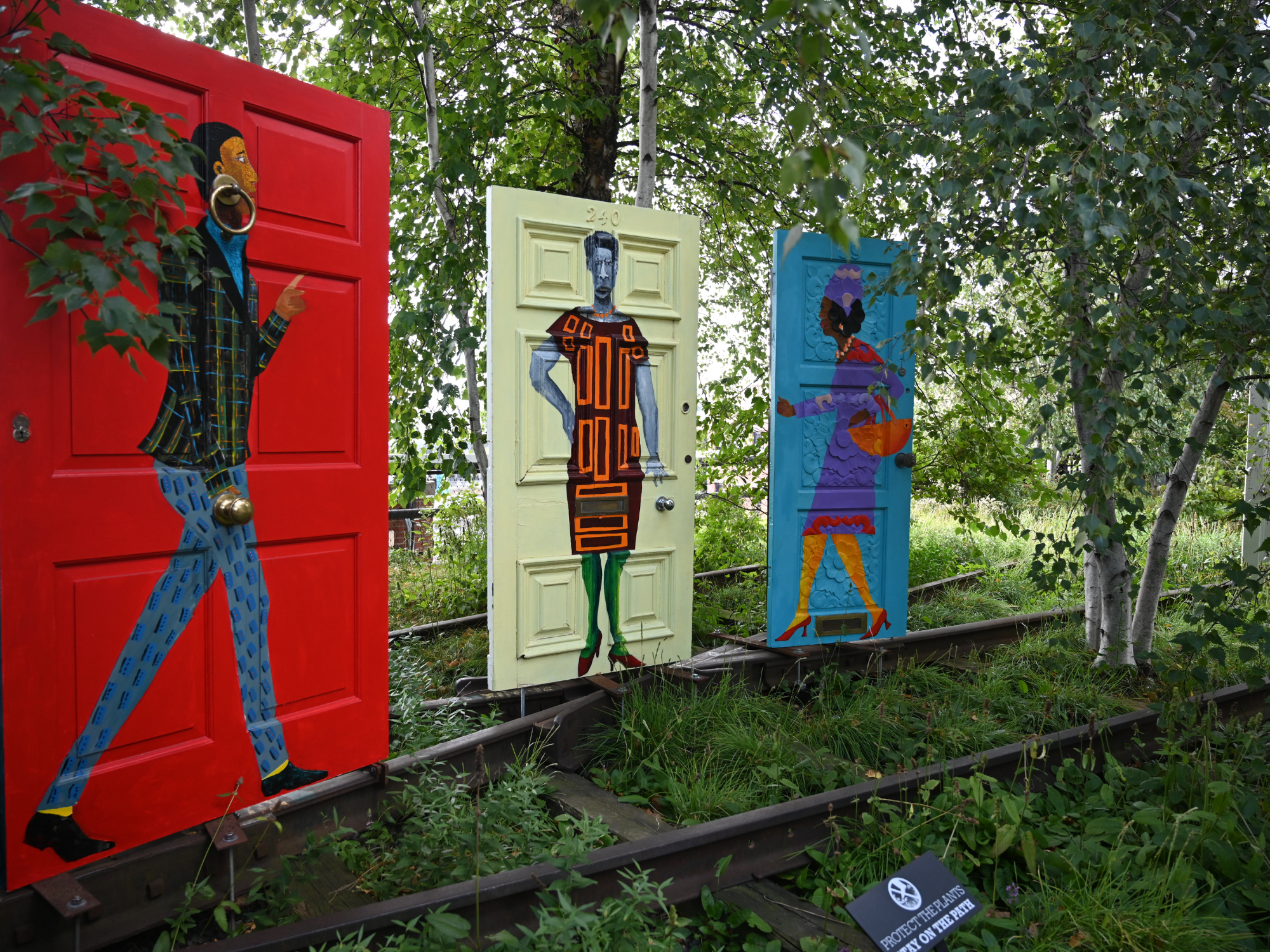 Three colorful doors stand on a grassy area, each featuring unique artistic designs of figures.