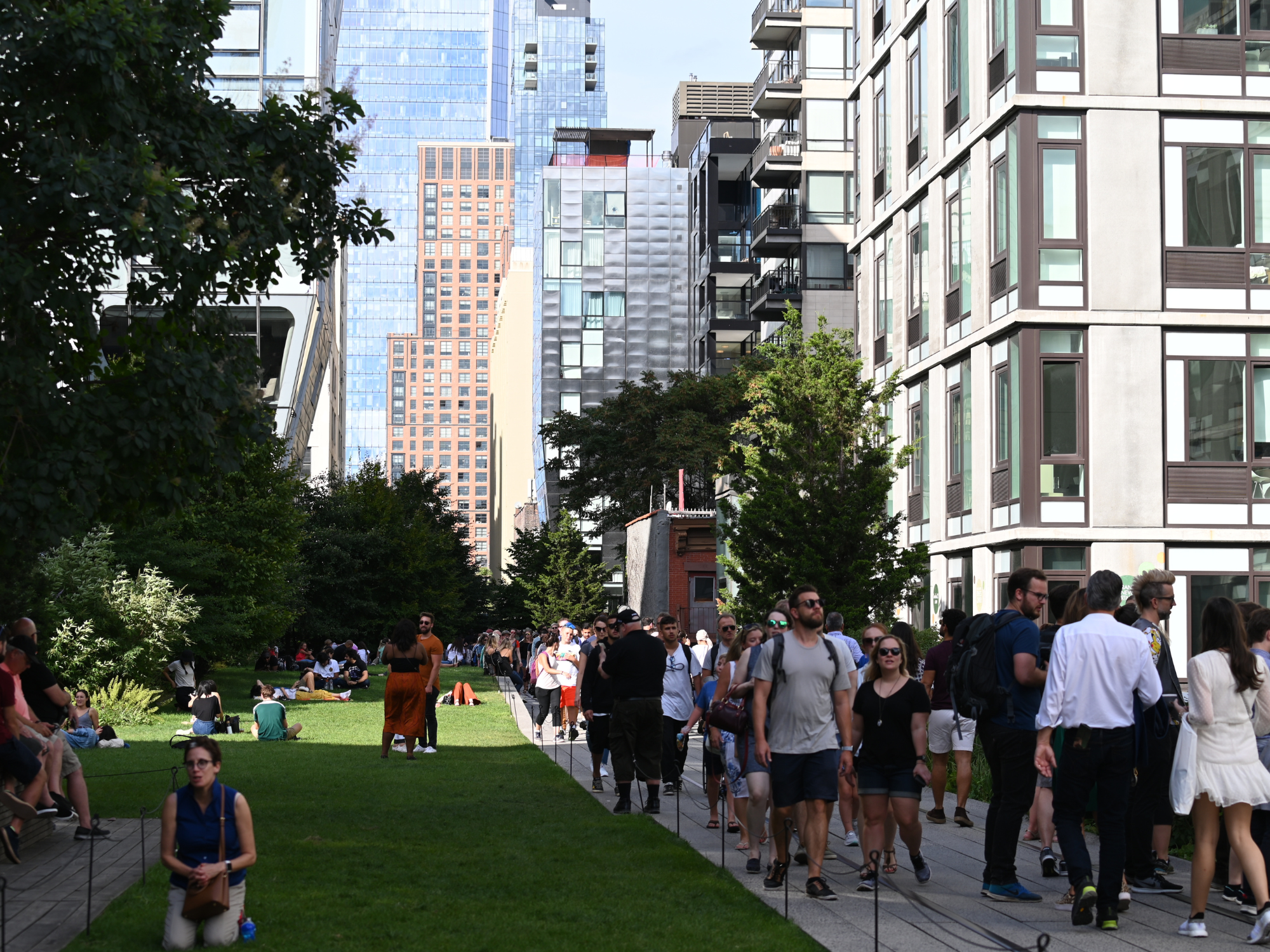 Crowd of people walking along a pathway in a park with modern buildings and greenery in New York.