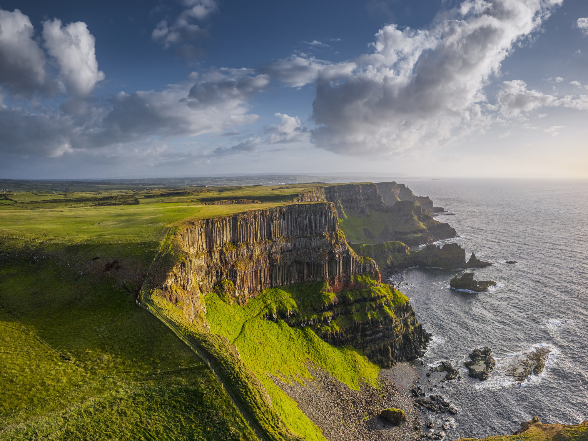 Cliffs overlooking the ocean with green fields and dramatic clouds in the sky, depicting a scenic Irish landscape.