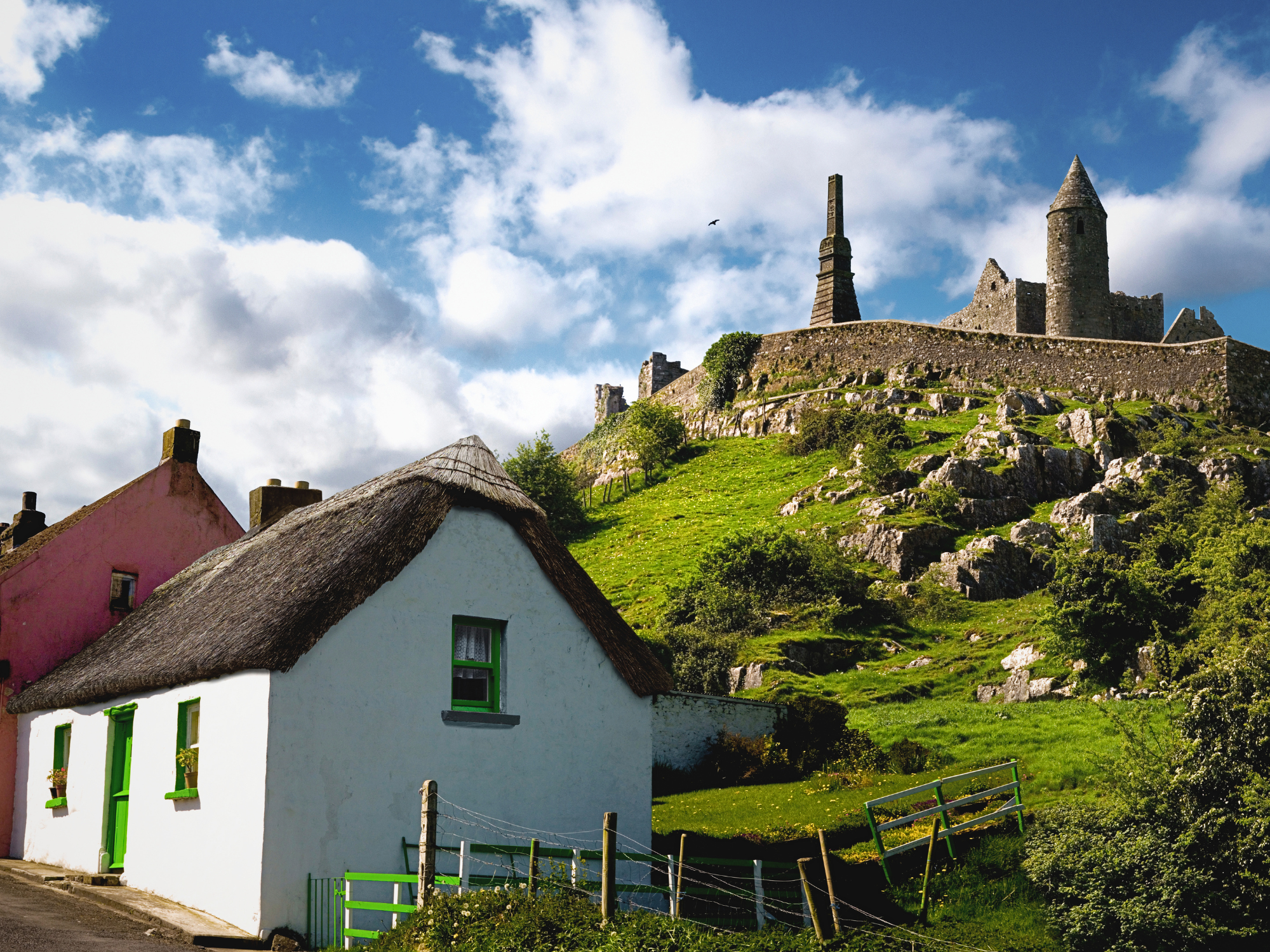 Thatched cottage in foreground with green door, stone castle on hill under blue sky with clouds, Ireland.