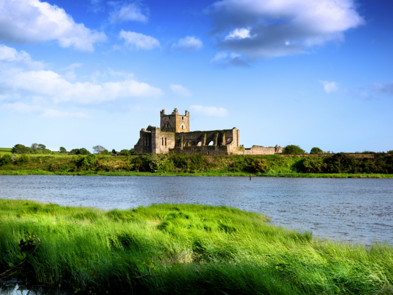 Ruins of a stone church beside a river, surrounded by lush green grass under a blue sky with clouds.