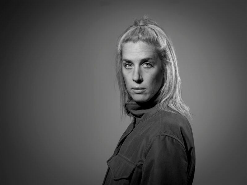 Black and white portrait of a woman with long, light hair, wearing a collared jacket, looking directly at the camera.