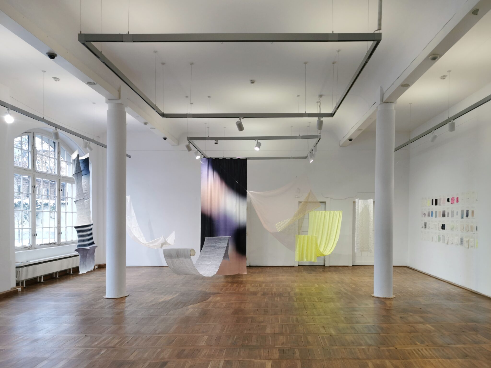 Art installation featuring flowing textiles suspended in a spacious gallery with wooden flooring and large windows.