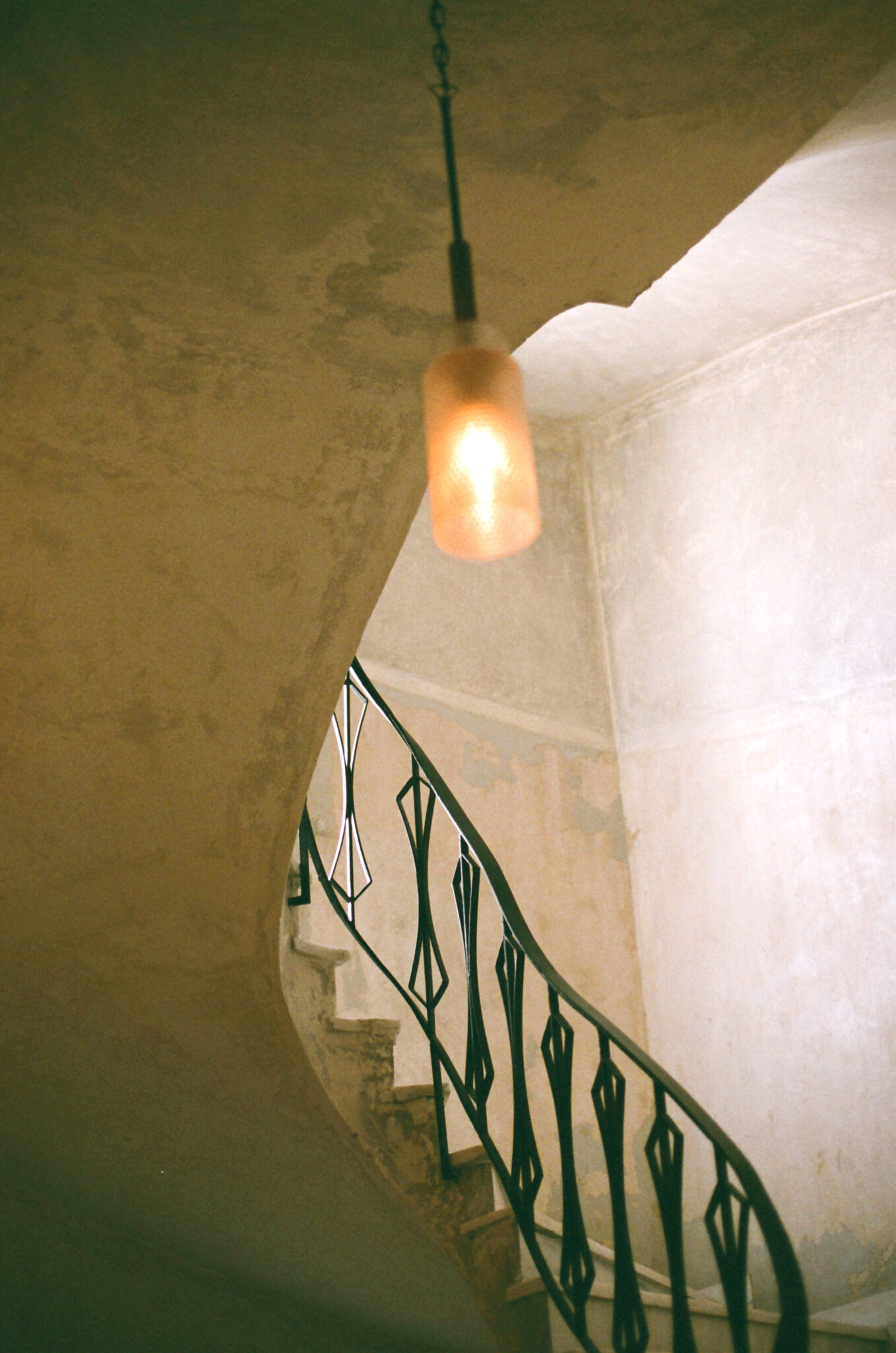Curved staircase with a decorative black railing and a warm pendant light, set against a textured wall.