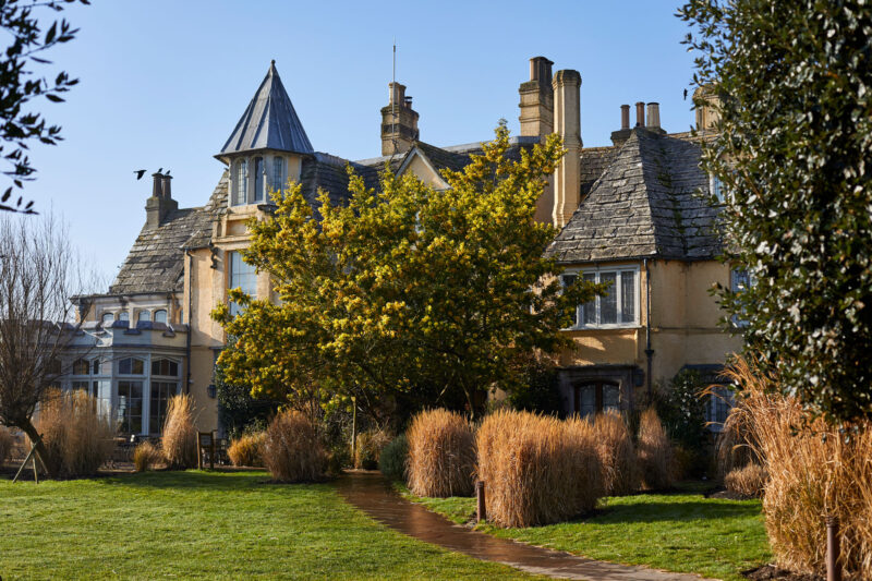Historic building with a pointed roof, surrounded by lush greenery and tall grasses, under a clear blue sky.