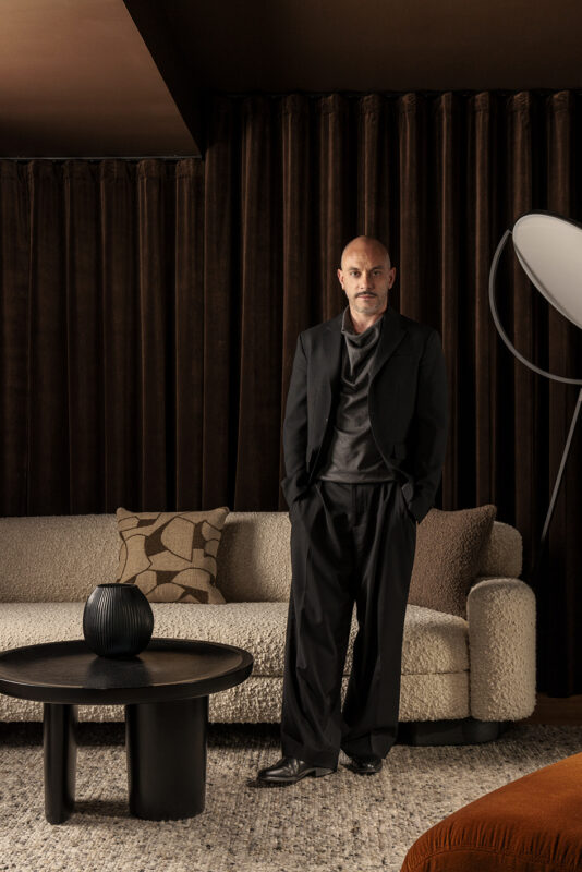 Fabien Louvier stands in a stylish interior, wearing a black suit and relaxed posture, beside a textured sofa and round ta...
