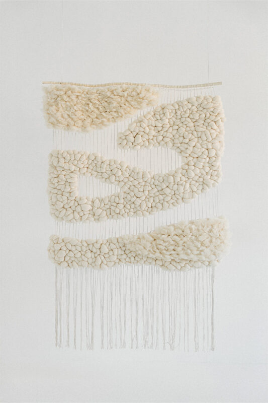 Textured wall hanging made of cream-colored stones arranged in flowing patterns, with hanging threads below.