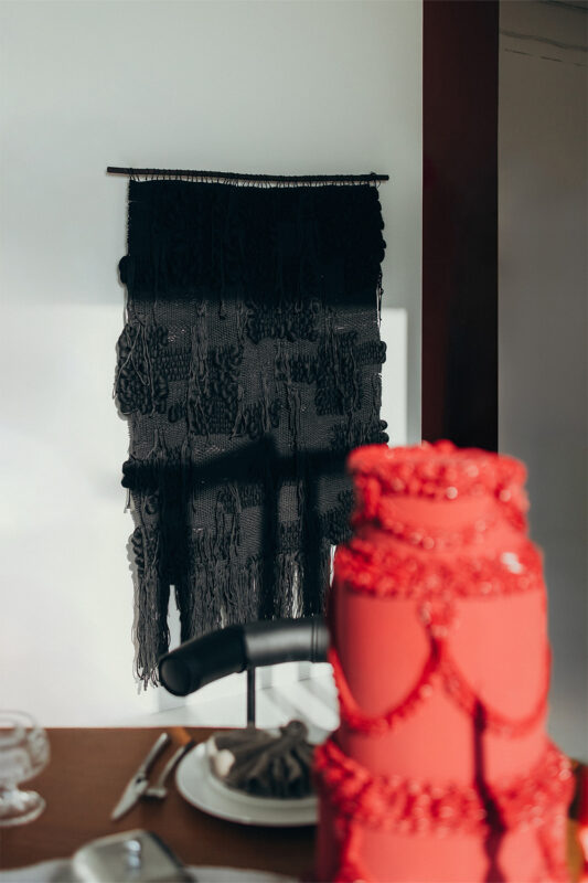 Black macramé wall hanging displayed in a bright room, with a red cake in the foreground.
