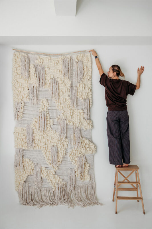 Person hanging a textured wall tapestry on a white wall while standing on a wooden stool.