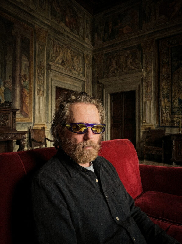 Man with a beard and sunglasses sits on a red couch in a richly decorated room with historical murals.