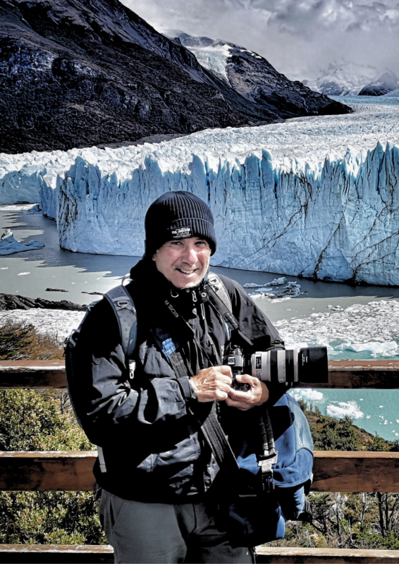 A person in a black jacket and beanie stands with a camera, overlooking a glacier and icy blue waters in a mountainous lan...