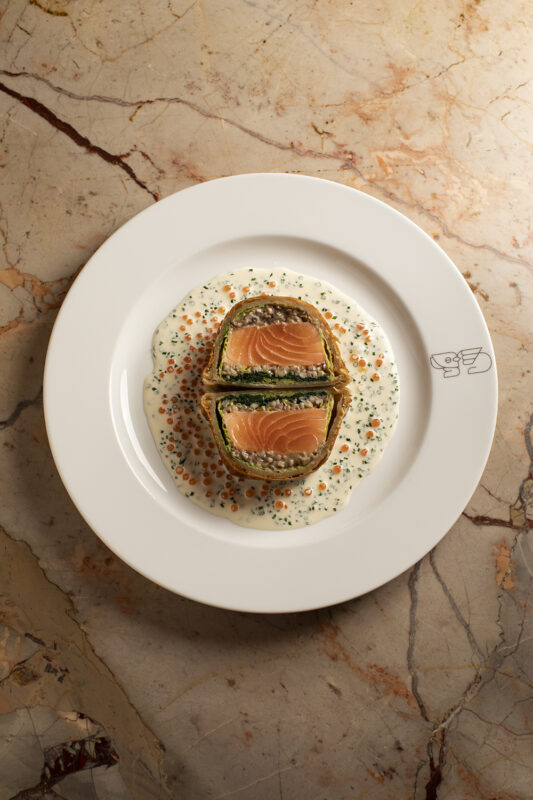 Coulibiac dish featuring layers of salmon and spinach, served on a creamy sauce with colorful caviar dots, on a decorative...