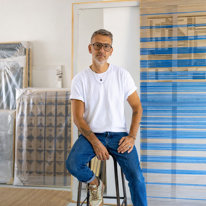 Man with glasses sitting on a stool in an art studio, wearing a white t-shirt and blue jeans, surrounded by artworks.