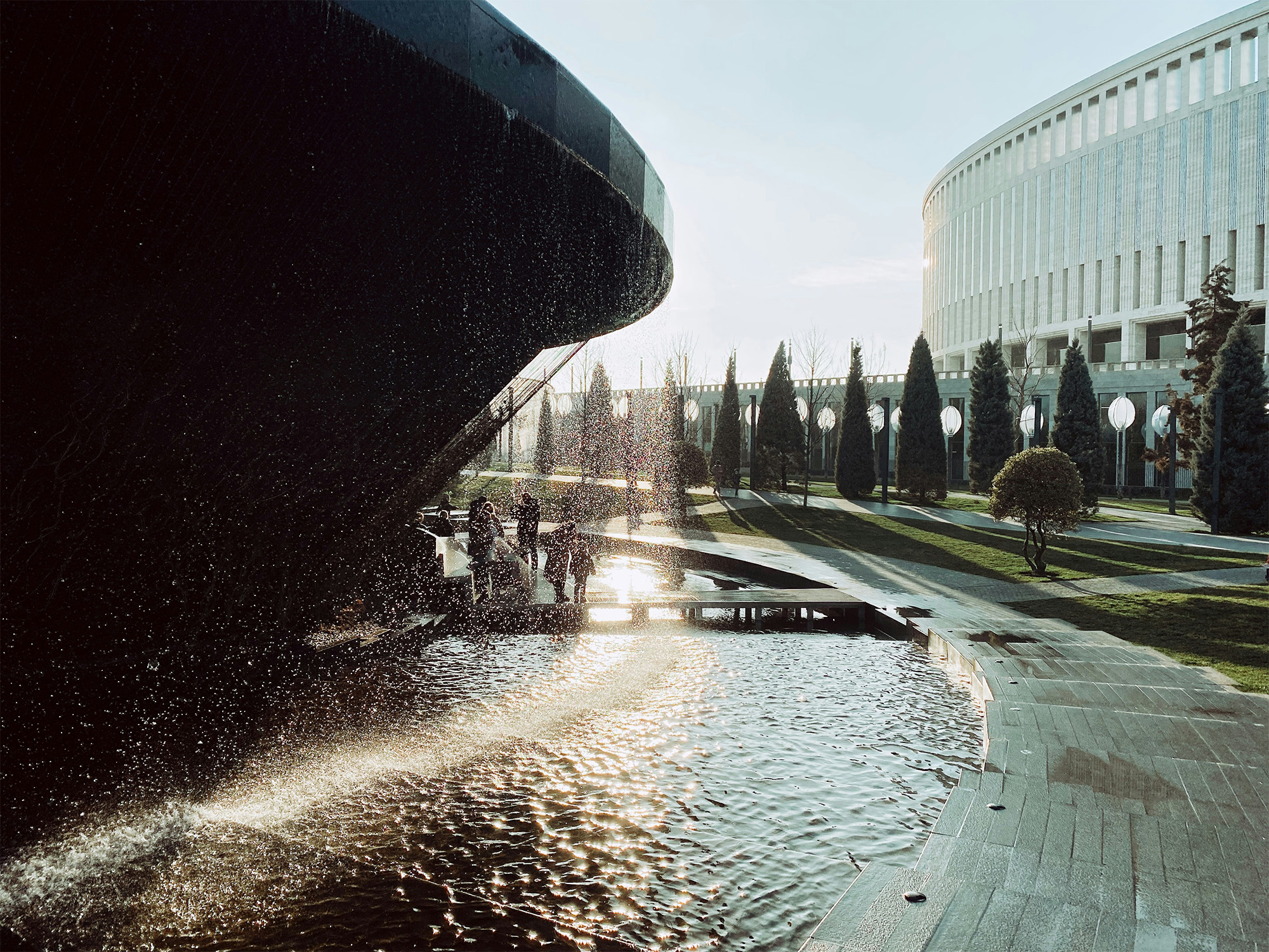 Fountain with water spray in a park, surrounded by trees and a modern building in the background.