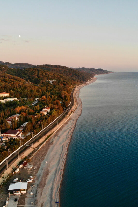 Aerial view of a coastal landscape with a sandy beach, railway tracks, and hills under a clear sky at sunset.