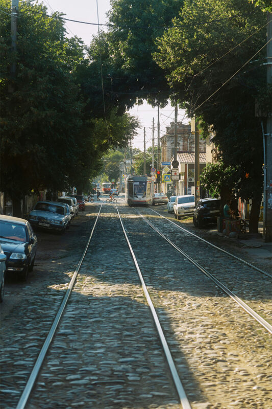 Cobblestone street with tram tracks, flanked by trees and parked cars, leading towards a distant tram in a sunny setting.