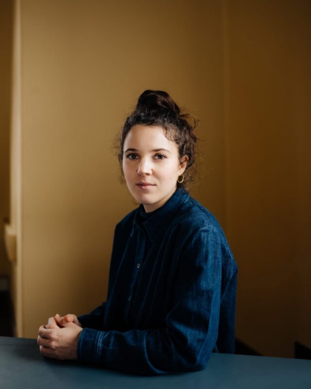 Portrait of a young woman with curly hair, wearing a denim shirt, seated at a table against a warm yellow background.