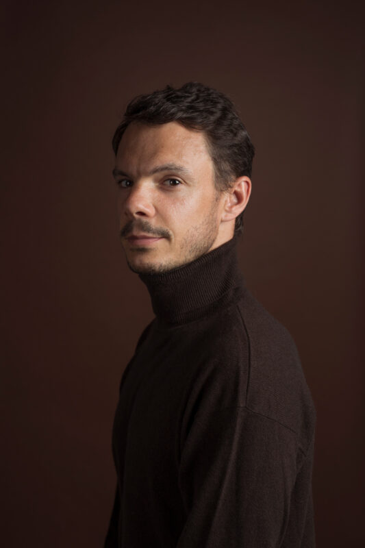 Portrait of a man with short dark hair, wearing a brown turtleneck, against a dark brown background.