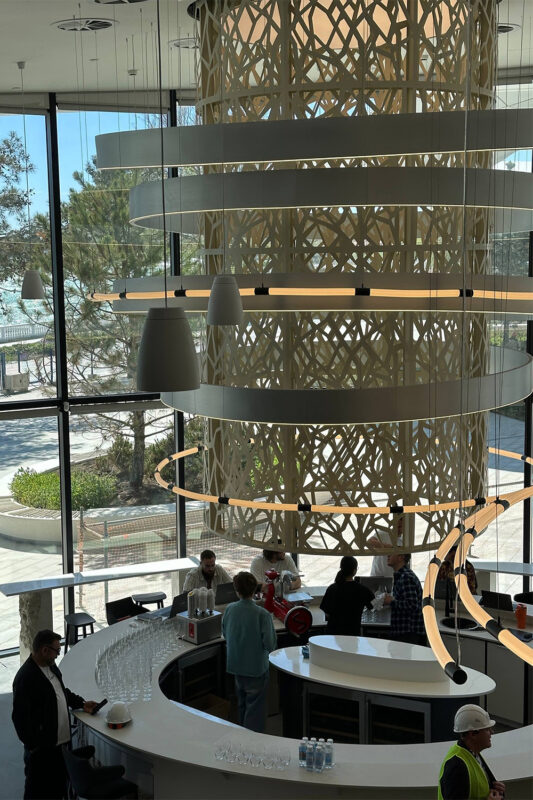 Modern interior of a café with a large decorative light fixture, patrons at the counter, and a view of greenery outside.