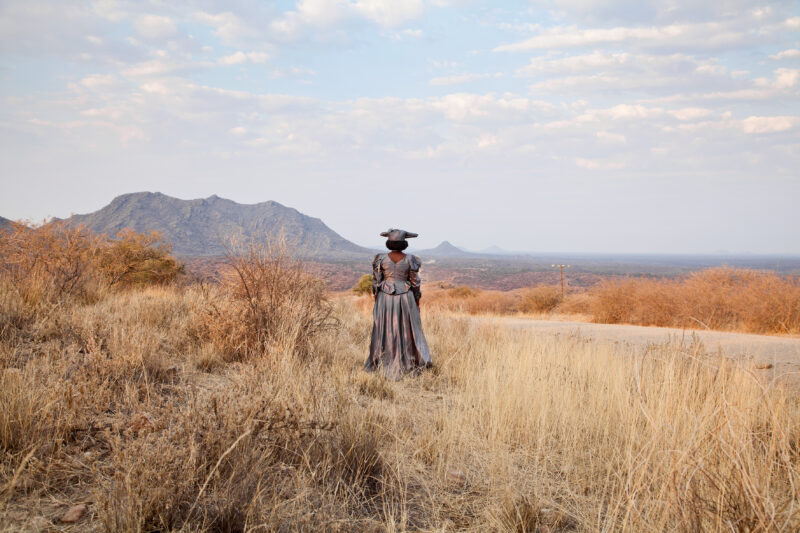 A person in traditional attire stands on a dirt road, gazing at distant mountains and a vast, dry landscape in Namibia.