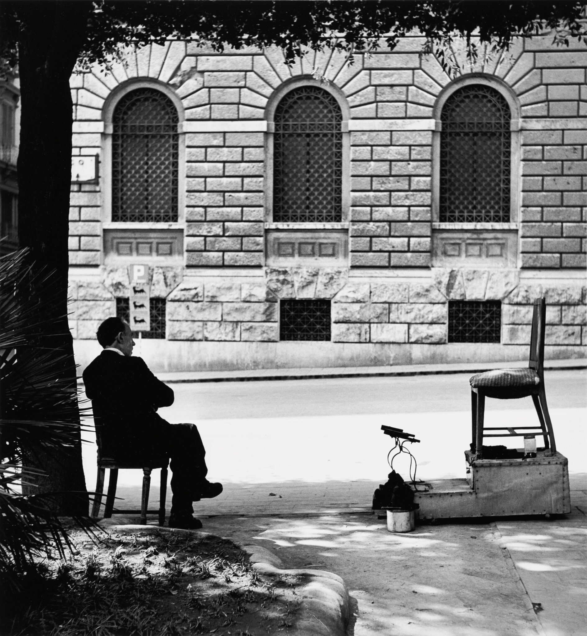 A man sits on a chair facing a street, with a shoe-shining station and a chair in front of him, framed by a stone building.