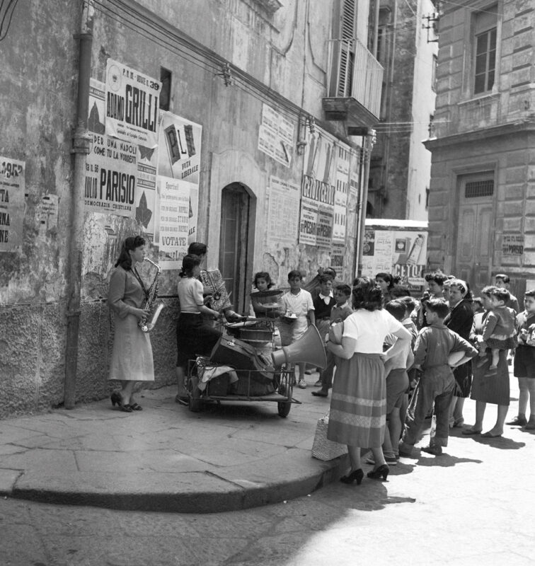 Children gather around a street performer playing saxophone in a historic urban setting, surrounded by posters.