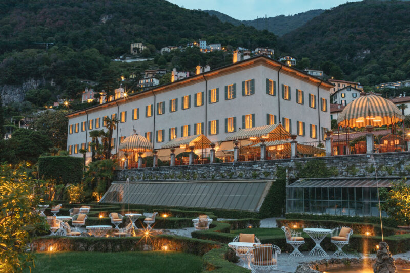 Elegant hotel exterior illuminated at night, surrounded by lush gardens and mountains in the background.