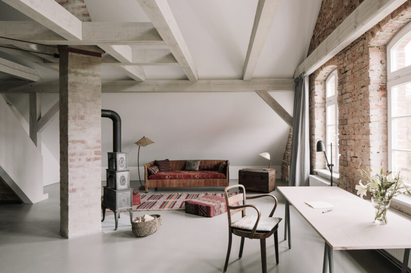 Stylish hotel room featuring a red sofa, wooden desk, and exposed brick walls, with natural light from large windows.