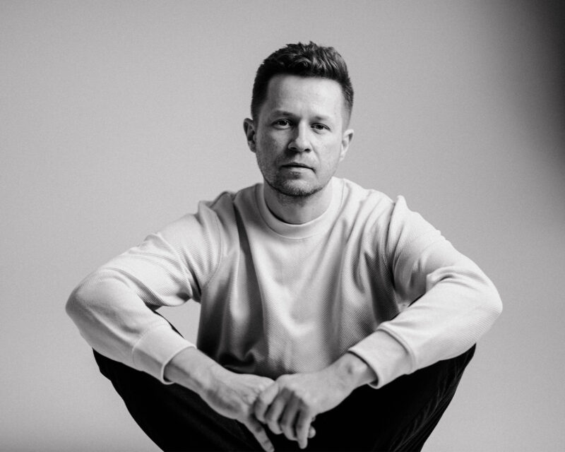 Black and white portrait of a man sitting on the floor, looking directly at the camera with a neutral expression.