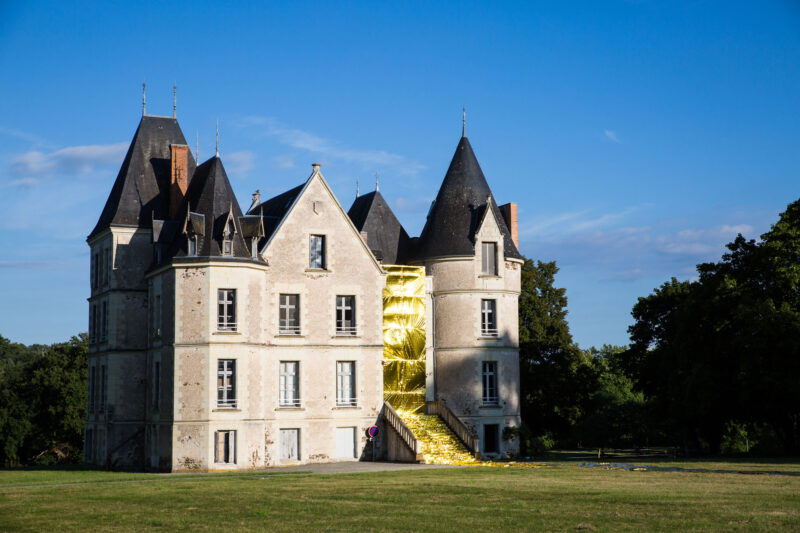 A historic chateau with a golden installation at its entrance, surrounded by green grass and blue sky.