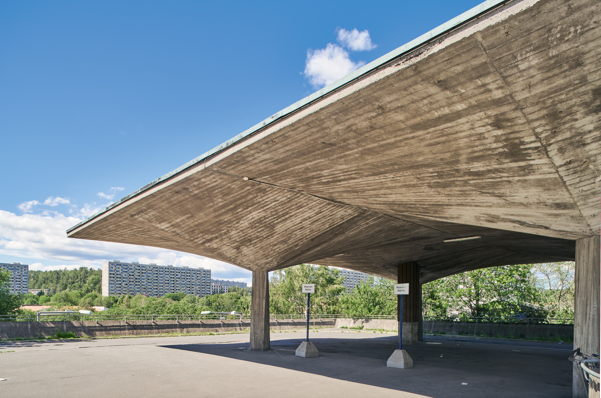 Concrete overhang with a curved design, casting shadows on a pavement, surrounded by greenery and residential buildings in...