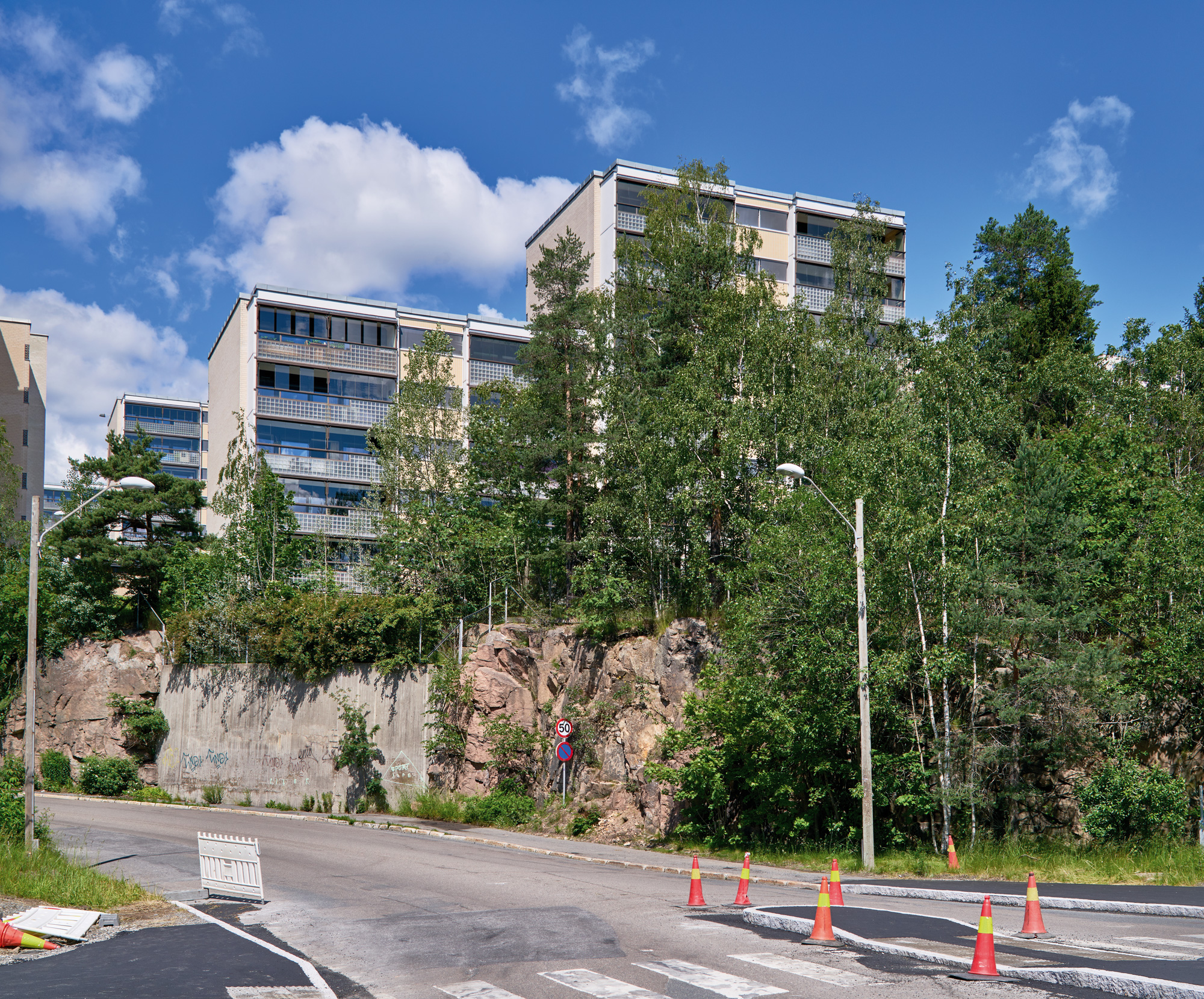 Modern buildings rise above a rocky outcrop, surrounded by trees, with a road and construction cones in the foreground.