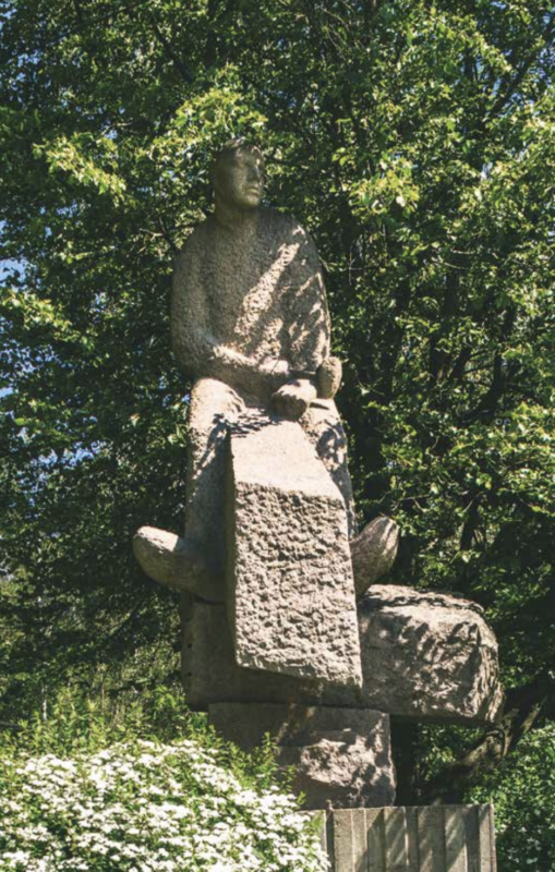 Stone statue of a seated figure atop a large block, surrounded by greenery and flowers in Oslo.