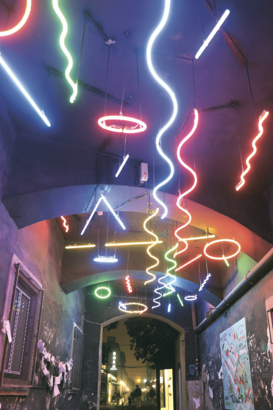 Colorful neon lights in various shapes hang from the ceiling of a dark corridor, creating a vibrant atmosphere.