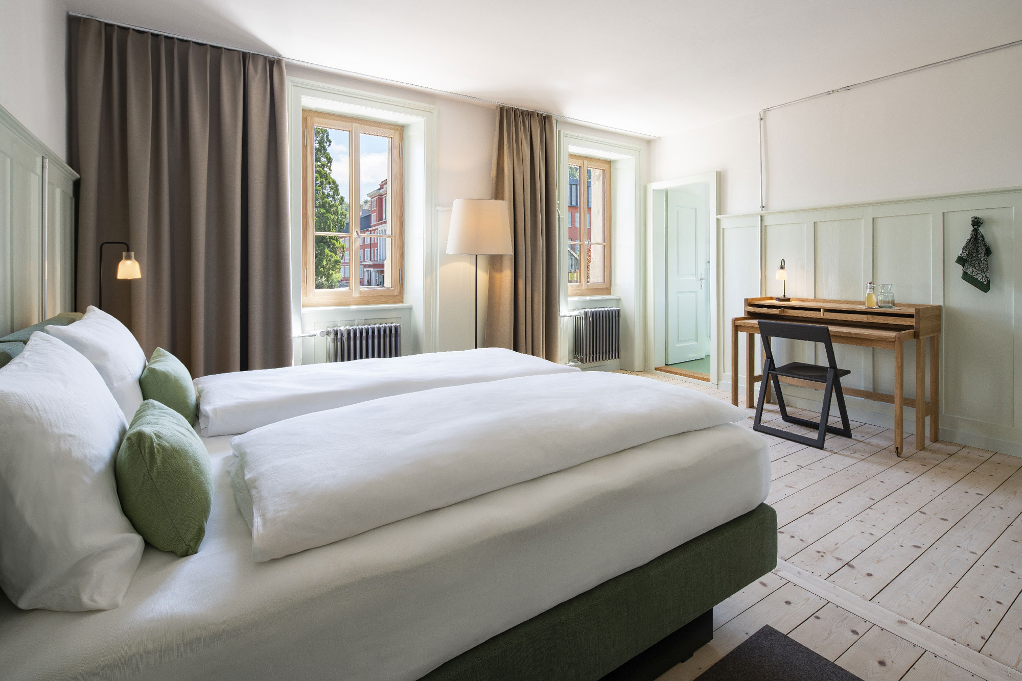 Modern guest room with two beds, green accents, large windows, and a wooden desk, featuring soft lighting and a cozy atmos...