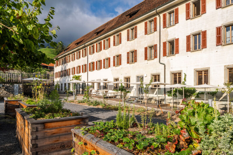 Historic building with red-shuttered windows, surrounded by garden beds and outdoor seating at Culinarium Alpinum.