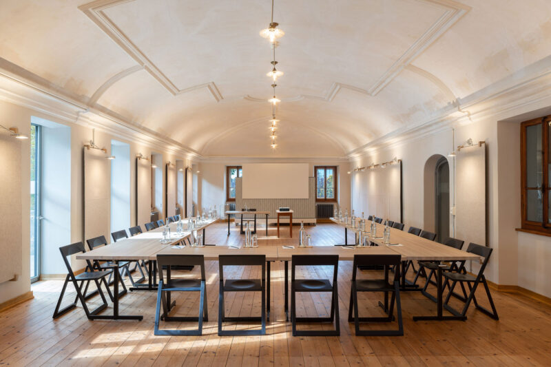 Modern conference room with a long table set in a U-shape, surrounded by black folding chairs, wooden floor, and large win...